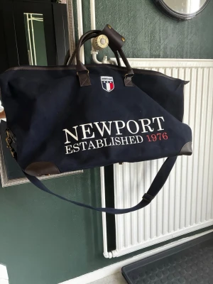 Marinblå weekendbag från Newport - Snygg marinblå weekendbag från Newport med stort vitt tryck 'NEWPORT ESTABLISHED 1976' och klubbmärke framtill. Väskan har bruna detaljer i läderimitation, dubbla handtag och justerbar axelrem. Perfekt för resor och övernattningar. Måtten på väskan är 77 cm lång x 42 cm hög x 35 cm bred.
