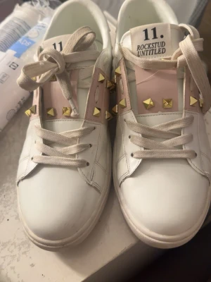 Valentino Rockstud Untitled sneakers - Snygga vita Valentino Rockstud Untitled sneakers med rosa detaljer och guldiga nitar. Skorna har klassisk låg profil, vita snören och är tillverkade i läder. Perfekta för dig som vill ha en unik och trendig look.