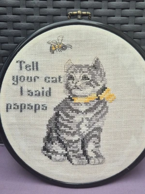 Tavla med busigt broderi  - Handgjord broderad tavla i rund svart ram med motiv av en grå katt med gul rosett och en bi. Texten 'Tell your cat I said pspsps' är broderad i grått. Tavlan är sydd på beige tyg och passar perfekt som dekoration för kattälskare.