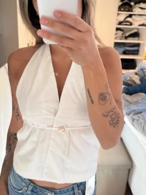 Vit halterneck topp med knyt - Säljer en snygg vit halterneck topp med knyt i midjan. Toppen har djup v-ringning och är ärmlös, perfekt till jeans eller kjol. Materialet känns som bomull och toppen har en croppad passform.