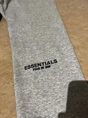 Gråa Essentials Fear of God mjukisbyxor - Snygga gråa mjukisbyxor från Essentials Fear of God. Byxorna har en enkel design med svart logga på benet och resår i midjan för skön passform. Tillverkade i mjukt material som passar perfekt för chill dagar.