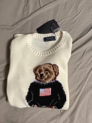 Vit stickad tröja med björn Polo Ralph Lauren - Vit stickad tröja från Polo Ralph Lauren med ikonisk björn på bröstet. Björnen har på sig en svart tröja med amerikansk flagga och blå jeans. Tröjan har rund halsringning och ribbade muddar. Perfekt för dig som gillar klassisk och lekfull stil.