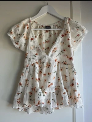 Blommig vit blus från Gina Tricot - Söt vit blus från Gina Tricot med orange och grönt blommönster. Blusen har korta ärmar, v-ringning och volangkant nedtill. Tillverkad i ett lätt, plisserat tyg som ger en luftig känsla. Perfekt för en somrig och avslappnad stil. Aldrig använt!