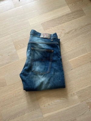 Blå slim fit jeans med slitningar - Säljer ett par blå slim fit jeans med snygga slitningar och tvättade detaljer. Jeansen har klassisk femficksdesign, normal midja och smala ben. Materialet är denim i bomull och de har en patch bak i midjan. W33 L32