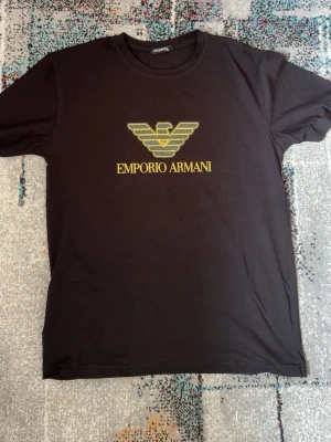 Svart Emporio Armani t-shirt - Snygg svart t-shirt från Emporio Armani med gul logga och text på bröstet. Klassisk rund hals och korta ärmar. Perfekt för dig som vill ha en stilren och trendig look med exklusiv känsla.