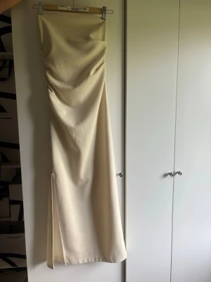 Lång beige bandeau klänning - Elegant långklänning i beige med axelbandslös design och draperad detalj på sidan. Klänningen har en slits på sidan  och en smickrande, figurnära passform. Perfekt för dig som vill ha en stilren och modern look. Använd fåtal gånger