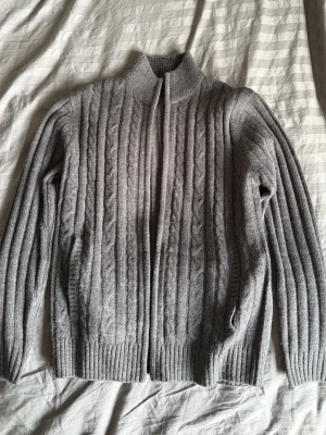 Grå kabelstickad kofta Stormy Life - Snygg grå kabelstickad kofta från Stormy Life med dragkedja framtill och hög krage. Koftan har långa ärmar, ribbade muddar och ett klassiskt mönster som ger en stilren look. Perfekt för lager på lager under kyliga dagar.