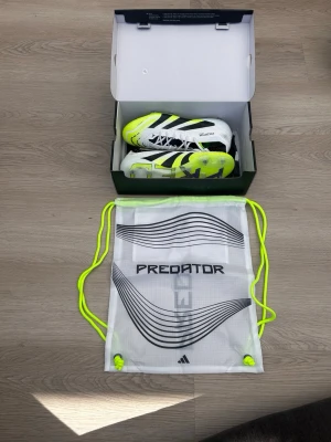Adidas Predator vita/gula sneakers - Säljer ett par Adidas Predator sneakers i vitt, svart och neon-gult. Skorna har klassiska svarta ränder, snörning och en sportig design med hög komfort. Ovandelen är i syntetmaterial och sulan är neonfärgad. Medföljer en vit och svart Predator-påse.