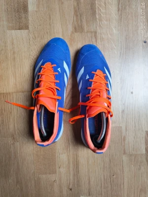 Blåa Adidas Predator fotbollsskor - Säljer ett par blåa Adidas Predator fotbollsskor med vita detaljer och orange snörning. Skorna har en låg profil och är tillverkade i syntetmaterial med gummisula som passar perfekt för inomhusspel. Snygg design och riktigt sköna på foten.