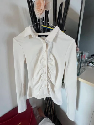 Vit skjorta med rynk från BikBok - Säljer en vit långärmad skjorta/blus från BikBok i storlek XS. Skjortan har knappar framtill och ett snyggt rynkat parti längs knappraden som ger en trendig look. Klassisk krage och figurnära passform. Perfekt till jeans eller kjol!