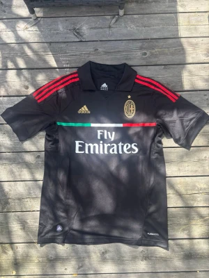 Svart AC Milan tröja från Adidas Oanvänd - Säljer en svart AC Milan matchtröja från Adidas med röda ränder på axlarna, klubbmärke och Adidas-logga i guld, samt Fly Emirates-tryck på bröstet. Tröjan har krage och detaljer i italienska flaggans färger. Perfekt för fotbollsfans! storlek L oanvänd 