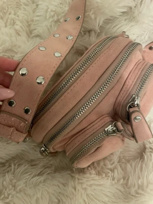 Ljusrosa mocka handväska - Säljer en ljusrosa handväska i mocka med flera dragkedjefack och bred axelrem dekorerad med silvriga nitar. Väskan har rundad form och detaljer i silverfärgad metall. Perfekt för dig som gillar en edgy men söt stil.