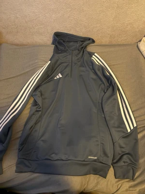 Mörkblå Adidas hoodie med dragkedja - Snygg mörkblå hoodie från Adidas med vit logga och klassiska tre ränder längs ärmarna. Tröjan har halv dragkedja framtill, huva och muddar vid ärmslut och nederkant. Tillverkad i funktionsmaterial med AEROREADY-teknologi.