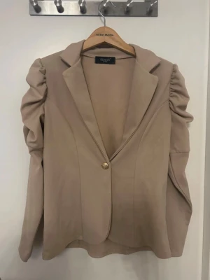 Beige kavaj med puffärmar - Stilren beige kavaj med markerade puffärmar och en knapp framtill. Klassisk krage och figurnära passform ger en trendig siluett. Perfekt för dig som vill sticka ut med en cool twist på den klassiska kavajen.