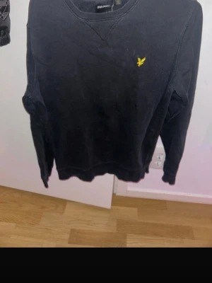 Svart crewneck - Klassisk svart sweatshirt från Lyle & Scott med den ikoniska gula loggan broderad på bröstet. Tröjan har rund halsringning, långa ärmar och ribbade muddar vid ärmslut och nederkant. Perfekt för en avslappnad och stilren look.