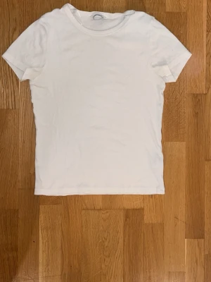 Vit basic t-shirt i bomull - En klassisk vit t-shirt i mjuk bomull med rund hals och korta ärmar. Perfekt att matcha med jeans eller under en skjorta för en clean och enkel stil.