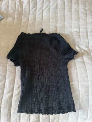 Svart smockad topp från H&M Divided - Svart smockad topp från H&M Divided med korta ärmar och vågig kant vid hals och nederdel. Toppen är stretchig och har en figurnära passform som ger en trendig look. Perfekt att styla med jeans eller kjol.