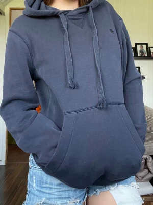 Mörkblå hoodie från G-Star RAW - Snygg mörkblå hoodie från G-Star RAW med klassisk huva och dragsnören. Tillverkad i mjukt material och har ribbade muddar vid ärmslut och nederkant. Liten G-Star-logga på bröstet för en clean look. Perfekt för en avslappnad stil.