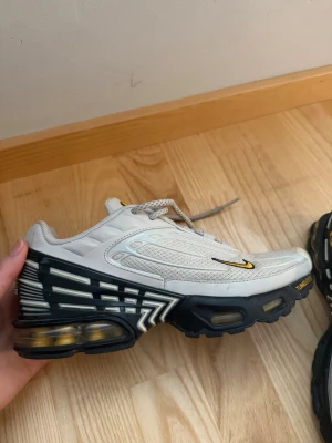 Nike air Max plus 3 - Nike Tuned Air sneakers i begie och svart med guldfärgade detaljer. Skon har en chunky sula med synliga luftkuddar och meshpaneler för extra ventilation. Snyggt mönster på sulan och liten guldfärgad Swoosh på sidan. Perfekt för dig som gillar streetwear.