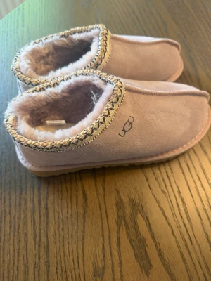 UGG tofflor Dam - Mysiga ljusrosa tofflor från UGG med mjukt fuskpälsfoder och dekorativ kant i beige och svart. Tofflorna har en klassisk slip-on modell och är perfekta för att hålla fötterna varma hemma. Utsidan är i mockaliknande material och sulan är lätt räfflad.