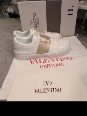 Valentino vita sneakers med gulddetalj - Kopior av vita sneakers från Valentino Garavani med bred guldfärgad rem över vristen och matchande guldfärgade nitar. Skorna är låga och har klassisk snörning samt detaljerad sula. Perfekta för dig som vill ha en lyxig touch på din outfit.