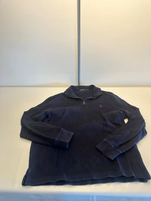 Mörkblå halvzip från Polo Ralph Lauren - Mörkblå långärmad tröja från Polo Ralph Lauren med halvzip och ståkrage. Tröjan har en liten röd broderad logga på bröstet och är tillverkad i mjukt material. Perfekt för en avslappnad men stilren look.