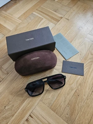 Tom Ford Falconer 02 pilotbågar solglasögon - Snygga svarta solglasögon från Tom Ford Falconer 02 med klassisk pilotform och mörka gråtonade glas. Ramen är i blank svart plast med ikoniska T-detaljer vid gångjärnen. Kommer med originalfodral och box. Perfekta för dig som vill ha en stilren och trendig look.