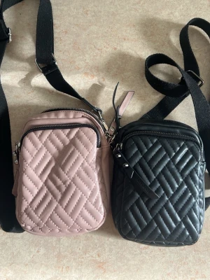 Två don donna axelväskor i rosa & svart - Säljer två små don Donna axelväskor i quiltad design, en i ljusrosa och en i svart. Båda har justerbara svarta axelband, dragkedja upptill och ett extra fack framtill. Perfekta för mobilen och småprylar. Materialet är konstläder med mjuk känsla.