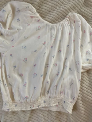 Vit blommig blus med puffärm - Supersöt vit blus med små blommor i rosa och blått. Blusen har puffiga korta ärmar och resår i midjan för en skön passform. Perfekt för dig som gillar romantisk och feminin stil.