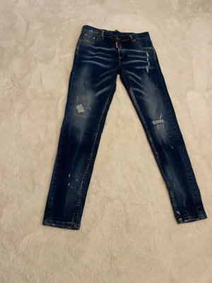 Mörkblå dsquared2 jeans med slitningar - Säljer ett par mörkblå dsquared2 jeans med coola slitningar och detaljer framtill. Jeansen har klassisk femficksmodell, smal passform och är tillverkade i denim. Perfekta för dig som gillar en trendig och avslappnad stil.