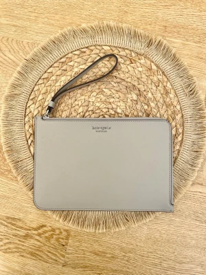 Grå kuvertväska från Kate Spade - Stilren grå kuvertväska från Kate Spade New York i slätt läder. Väskan har en avtagbar handledsrem och minimalistisk design med silverfärgad logga framtill. Perfekt för dig som gillar enkel och klassisk stil.