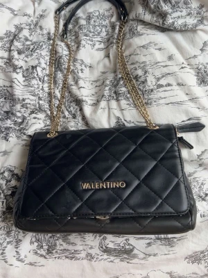 Svart quiltad handväska Valentino - Snygg svart handväska från Valentino med quiltat mönster och guldfärgade kedjehandtag. Väskan har en klassisk rektangulär form och dragkedja på sidan. Perfekt accessoar för att lyfta din outfit.