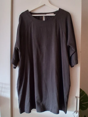 Mörkgrå loose klänning Rodebjer  - En enkel och stilren mörkgrå klänning i loose fit med trekvartsärmar och rund halsringning. Klänningen har en öppning med knapp i nacken och är tillverkad i ett mjukt tyg, perfekt för en avslappnad look.