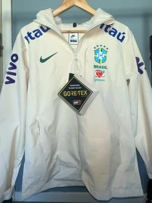 Vit Nike Brasil vindjacka Gore-Tex - Snygg vit vindjacka från Nike med Brasil CBF-logga och sponsordetaljer i grönt, blått och rött. Jackan har huva, hel dragkedja och är tillverkad i slitstarkt Gore-Tex-material. Perfekt för dig som gillar sportig stil och vill hålla dig torr.
