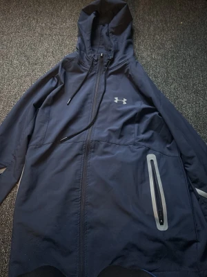 Marinblå vindjacka från Under Armour - Snygg marinblå vindjacka från Under Armour med huva och dragkedja framtill. Jackan har reflekterande detaljer och en praktisk ficka med dragkedja på sidan. Perfekt för blåsiga dagar och sportiga outfits.
