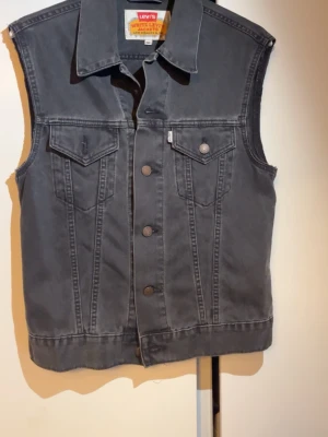 Svart Levis väst - Snygg svart ärmlös jeansjacka från Levi's med klassisk krage, två bröstfickor med lock och knappstängning framtill. Perfekt att slänga över en hoodie eller t-shirt för en cool och avslappnad look.
