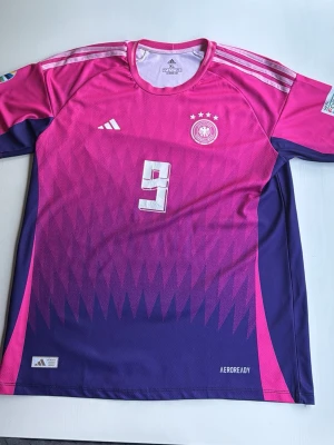 Tyskland EM tröja - Snygg Tyskland matchtröja från Adidas i rosa och lila med vita detaljer. Tröjan har nummer 9 och namnet Füllkrug på ryggen, samt fyra stjärnor ovanför landslagsemblemet. Tillverkad i lätt och ventilerande material med AEROREADY-teknologi.