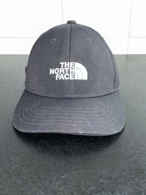 Svart keps från The North Face - Snygg svart keps från The North Face med klassisk böjd skärm och justerbart spänne bak. Kepsen har broderad vit logga framtill och är tillverkad i ett slitstarkt tyg som passar perfekt till streetwear-looken.