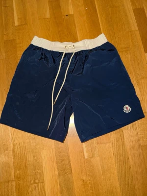 Marinblå badbyxor med vit midja - Snygga marinblå badbyxor från Moncler med vit resårmidja och dragsko. Klassisk passform, Moncler-logga på benet och en bakficka med knapp. Perfekta för stranden eller poolen. Materialet är snabbtorkande och lätt.