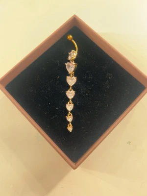 Guldig navelpiercing med hjärtformade stenar - Säljer en navelpiercing i guldton med flera hjärtformade stenar på. Rengör alltid mina smycken innan jag postar!💓💓