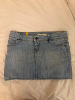 Low waist jeans kjol - Säljer en snygg ljusblå jeanskjol i klassisk modell med fem fickor, knapp och dragkedja framtill. Kjolen har guldfärgade detaljer och kontrastsömmar. Perfekt till en avslappnad och trendig look.