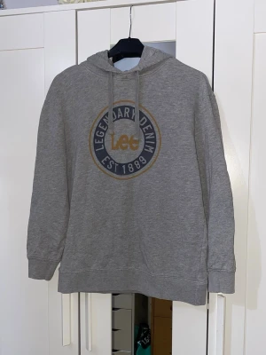 Grå hoodie från Lee med tryck - Snygg grå hoodie från Lee med stort tryck på bröstet där det står 'Legendary Denim Est. 1889'. Hoodien har huva med dragsko och mörkblå armbågslappar på ärmarna. Perfekt för en avslappnad och cool stil.