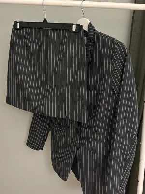 Randig kostym med kavaj och kjol - Säljer en mörkgrå kostym med vita ränder, består av en lång kavaj och en matchande kort kjol. Klassisk pinstripe-look som ger en cool och stilren vibe. Perfekt för dig som vill sticka ut med en trendig twist på klassikern.