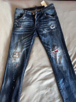 Mörkblå distressed jeans Dsquared2 - Säljer ett par mörkblå jeans från Dsquared2 med slitningar, hål och färgstänk för en edgy look. Jeansen har fem fickor, raka ben och coola detaljer både fram och bak. Perfekta för dig som gillar streetstyle och unika plagg.