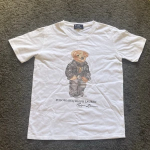 Ralph Lauren T-shirt  - Vit t-shirt från Ralph Lauren med ikoniskt Polobear-tryck framtill. Klassisk passform och rund halsringning. Tillverkad i mjuk bomull som känns skön mot huden. Perfekt för dig som gillar stilrena plagg med en twist. Priset kan diskuteras.