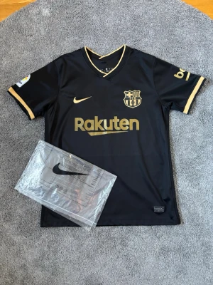 Svart FC Barcelona matchtröja  - Snygg svart FC Barcelona matchtröja från Nike med guldfärgade detaljer och loggor. Tröjan har klubbmärke, Nike-logga och Rakuten-tryck på bröstet samt guldfärgade kanter vid ärmar och krage. Perfekt för dig som älskar fotboll och vill sticka ut på plan.