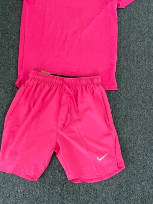 Nike träningsset  - Säljer ett par neonrosa träningssets från Nike med elastisk midja och snörning. Shortsen har standard fit och är tillverkade i ett lätt syntetmaterial, perfekta för sport eller gym. Nike-logga i vitt på ena benet för en sportig look.