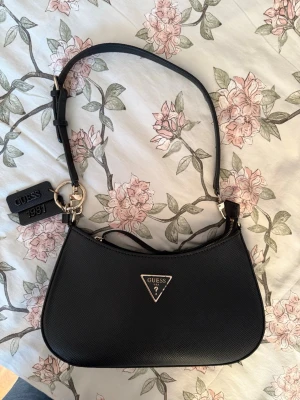 Svart axelremsväska från Guess - Snygg svart handväska från Guess med justerbar axelrem och guldfärgade detaljer. Väskan har Guess-logga i metall framtill och en tag med texten 'Guess 1981'. Tillverkad i ett strukturerat konstläder som ger en lyxig känsla. Väskan är i bra skick men har nötts lite på nyckelringarna som syns på bilderna samt att det är lite läppglans i innetyget i det stora facket