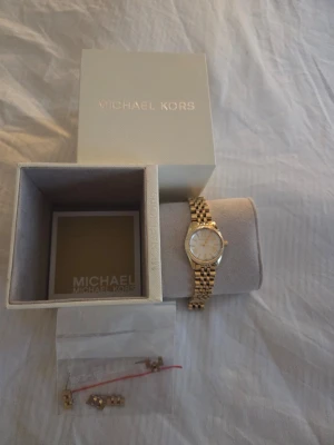 Guldfärgad klocka från Michael Kors - Stilren guldfärgad klocka från Michael Kors med klassisk länkarmband och romerska siffror på urtavlan. Klockan har en rund form och levereras med originalask samt extra länkar. Perfekt accessoar för att lyfta din outfit. Sparsamt anävnd unga repor eller skador.  Köpt för flera år sedan cirka för 2499kr