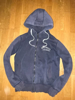 Mörkblå hoodie med dragkedja - Mörkblå hoodie från Race Marine med dragkedja framtill och vit text på bröstet. Utrustad med huva och vita dragsnören. Mjuk fleece på insidan för extra komfort. Två fickor framtill och ribbade muddar vid ärmar och nederkant.